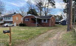 A charming single-story brick house at 103 W Thorndale Drive with a front porch overlooks a yard with a grass driveway. A black mailbox on a wooden post stands proudly, while trees and neighboring homes frame this Oxford gem in the background.