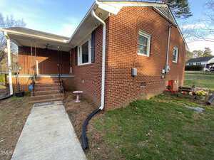 A one-story brick house with a small yard. The house has a side entrance with a few steps leading up to a porch. A concrete path runs alongside the house. A pink birdbath and some decorations are in the yard.