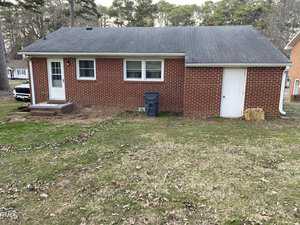 A small one-story red brick house with a gray shingled roof. The house has a white door and two windows. A trash bin is positioned against the back wall. The backyard is grassy with scattered leaves, surrounded by trees and neighboring houses.