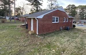 A red brick house with a gabled roof is situated on a grass lawn. The house has a small rectangular window and an air conditioning unit on one side. Trees and other houses are visible in the background.