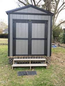 A gray storage shed with double doors and black trim stands in a grassy yard. There are three wooden steps leading up to the entrance, and lattice skirting surrounds the base. Trees and a fence are visible in the background.