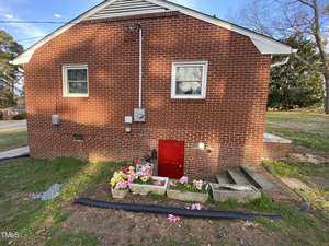 A red-brick building is shown with two small windows. A red door is at the base, accessed by a short set of stairs. In front, there are flower boxes with pink and white flowers. A black drainage pipe runs along the ground.