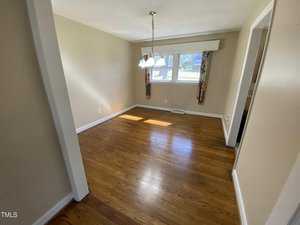 A small, empty room with beige walls and a wooden floor. A central light fixture hangs from the ceiling. Two windows with floral curtains allow natural light to enter. A doorway is visible on the right side of the room.