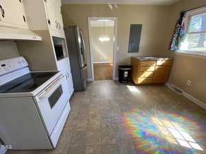 A kitchen with white appliances, including a stove and refrigerator. There is a wood cabinet near a window on the right. The floor is tiled, and light from the window creates a colorful reflection. A doorway leads to another room.
