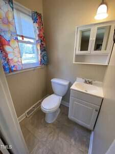 A small bathroom with a white toilet, white vanity with sink, mirrored medicine cabinet, beige walls, tiled floor, and a window with colorful floral curtains letting in natural light.