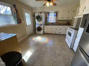 A kitchen with white cabinets, a stove, refrigerator, and stacked washer and dryer by the door. Sunlight creates a rainbow effect on the floor. Colorful curtains hang by the window and door.