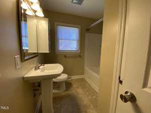 A bathroom with tan walls and tile floor, featuring a pedestal sink, toilet, wall mirror with lights, towel bar, and a bathtub with a shower and curved curtain rod. Two windows are above the toilet and tub.