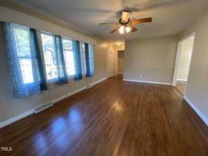A spacious room with polished hardwood floors and beige walls. A ceiling fan with lights is centrally mounted. Large windows with blue curtains allow natural light to enter. The room leads to a hallway with a white door.