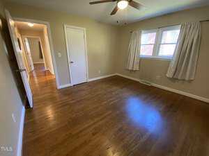 Empty room with tan walls, hardwood floors, a ceiling fan, a window with white curtains, two closed doors, and a view into a hallway with more hardwood flooring.