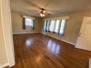 A spacious living room with polished hardwood floors and beige walls. A ceiling fan with lights is centered on the ceiling. Three windows with blue curtains let in natural light, and a white door is visible on the right.