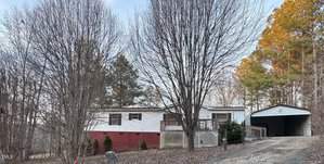 A single-story manufactured home at 328 Cone Drive features a porch, raised on a red foundation, surrounded by barren trees. A gravel path leads to an adjacent carport in Henderson. Pine trees stand tall in the background under a clear sky.