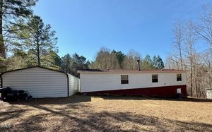 A single-story mobile home with a white upper section and red lower section sits on a grassy lot. A detached white garage is on the left. The area is surrounded by trees under a clear blue sky.