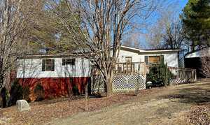 A single-story mobile home with white siding and a red foundation is surrounded by bare trees. There is a wooden deck attached to the front of the house. The yard is covered in dry leaves and a sloped gravel path leads to the entrance.