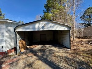 A small, open carport with a pitched roof is attached to a building. The area is surrounded by trees, and the ground is covered with gravel and patches of grass. A wooden board leans against the carport's left wall. The sky is clear and blue.