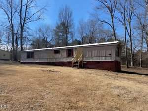 A long, single-story mobile home with beige siding and red shutters sits on a red brick foundation in a grassy yard on 415 Island Creek Estates Road, Henderson. Several leafless trees dot the landscape under the clear blue sky. Steps lead invitingly up to the front door.