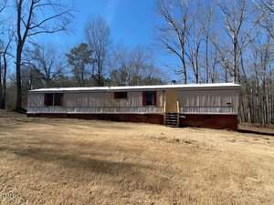 A single-story mobile home stands on a grassy plot with bare trees in the background. The home has a light exterior with red trim and several windows. A short staircase leads to the main entrance. The sky is clear and blue.