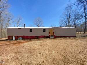 A long, rectangular mobile home with a red base and light gray upper section is situated in a rural area. It has a chimney and a set of red stairs leading to a yellow door. The surrounding area features dry grass and leafless trees under a clear blue sky.