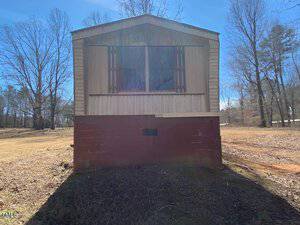 A mobile home with brown lower siding and tan upper siding sits on a grassy lot. There are trees without leaves in the background and clear blue sky above. The front windows have dark shutters, and the shadow of the home is visible on the ground.