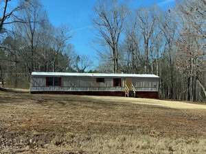 A long, single-story mobile home with a reddish-brown foundation and a wooden porch sits in a grassy yard. Bare trees without leaves surround the home, and a clear blue sky is overhead.