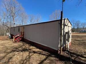 A long, narrow mobile home with light-colored siding and a red staircase sits in a yard. Bare trees and a clear blue sky are in the background. There's a utility pole nearby, and a small white house is visible in the distance.