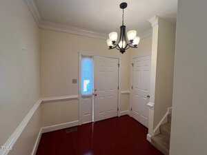 A small, empty foyer with hardwood floors, cream-colored walls, and white trim. There are three white doors, a small staircase with beige carpet, and a black chandelier with frosted glass shades hanging from the ceiling.