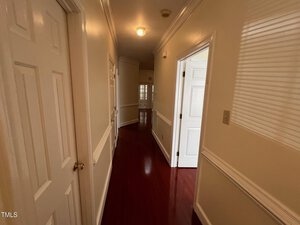 A hallway with polished wooden floors, cream-colored walls, and white doors. The ceiling has a light fixture, and molding decorates the top and middle of the walls. One door is partially open, leading to another room with a window.