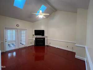 A living room with high ceilings featuring a ceiling fan and two skylights. A fireplace with a black surround is centered on the far wall, with a flat-screen TV above it. Double doors with glass panes lead to another room with a checkered floor.