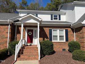 The brick house at 2002 Fernwood Court Unit 2, Henderson, boasts a white porch and a red front door adorned with a wreath. With white trim and dark shutters, steps lead to the entrance, flanked by green bushes. Majestic trees stand in the background under a clear blue sky.