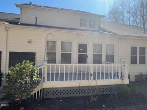 A white wooden house with a small raised deck, featuring railing and two hanging lanterns. There are multiple windows, a door on the left, and a sloping roof. A bush is on the left, and bare trees are in the background under a clear blue sky.