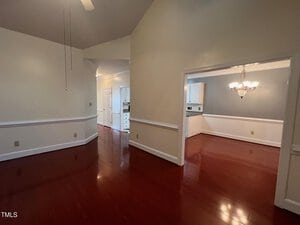The image shows an empty interior space with wooden floors and cream-colored walls. There are two rooms visible, one with a chandelier and a gray accent wall. A hallway leads to another area with white cabinets. A ceiling fan is partially visible.