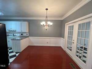 A kitchen with white cabinets, a black and white checkered floor, and a dark countertop is visible. Adjacent is a dining area with red hardwood flooring, a chandelier, and double glass-paneled doors leading to another room.