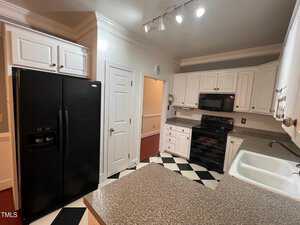 A kitchen with black and white checkered floor, white cabinets, and dark countertops. Black appliances include a refrigerator, oven, and microwave. There's a white double sink and ceiling track lighting above. A doorway leads to another room.
