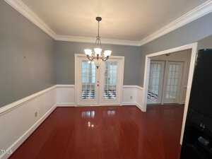 A dining room with wooden flooring and gray walls, featuring a chandelier hanging from the ceiling. Double French doors open to another room with black and white checkered flooring. A portion of a black refrigerator is visible on the right.