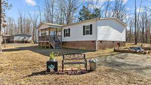 A white manufactured home with a brick foundation is set in a wooded area. A front porch with steps leads to the entrance. In the foreground, there's a black metal bench, a potted plant, and a small raised garden bed. Sparse trees surround the area.