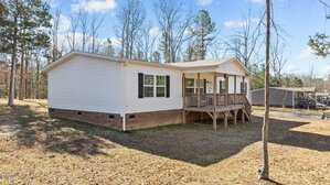 A single-story manufactured home with light-colored siding and a brick foundation sits on a grassy lot with sparse trees. The home features a small front porch with a railing and steps leading to the front door. The sky is clear and sunny.