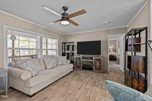 A living room featuring a beige sofa with patterned cushions on a wooden floor. A large flat-screen TV is mounted on a stand with cabinets. The room has a ceiling fan, two tall shelves, and large windows with blinds on the left. A doorway leads to another room.
