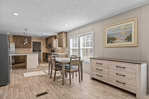 A modern kitchen and dining area with light wood flooring and cabinetry. The room features a dining table with four chairs, a large island, stainless steel appliances, and a landscape painting on the wall. Sunlight filters through the window blinds.