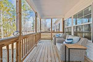 Front porch with wooden flooring and railing, featuring two wicker chairs with cushions and a small wooden side table. The porch is covered with a white ceiling, and the background shows trees and a nearby building.