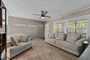 The image shows a living room with two beige sofas facing each other. There are decorative pillows on each sofa. A ceiling fan with a light fixture is centered overhead. Large windows with blinds are on the right wall, and the floor is wood-patterned.