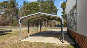 A metal carport with a curved roof stands next to a brick and siding building. It covers a concrete slab. In the background, a road is visible with a truck driving by, and there are trees lining the roadside.