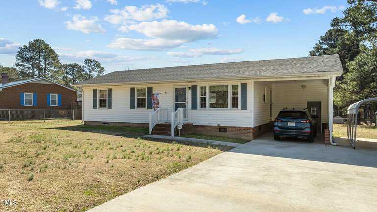 A single-story white house on 1397 Ruin Creek Road boasts a brick foundation and a small porch. The driveway leads to a carport on the right with a parked car. The lawn is sparse, with some grass patches, and the neighboring Henderson home is partially visible on the left.