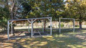 A wooden picnic table is under a simple open-sided shelter with a slanted roof in a fenced area. The structure is surrounded by trees, and sunlight filters through the branches, casting shadows on the grass.