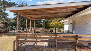 A wooden deck with a partial roof is attached to a white house. The deck has a railing and several patio chairs. The area is surrounded by trees and grass under a partly cloudy sky.