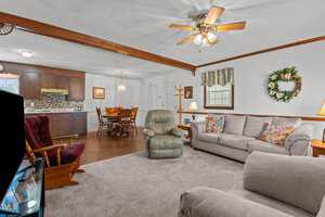 A cozy living room and dining area with a ceiling fan. The space includes a sofa, recliner, dining table with chairs, and a kitchen area with wooden cabinets. Decor includes a wall wreath, floral pillows, and a patterned curtain.