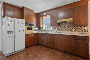 A vintage kitchen with dark wooden cabinets and a brown tile backsplash. Features include a white refrigerator with magnets, a yellow oven and range hood, a window above the sink, and a microwave on the counter. The floor is wooden.