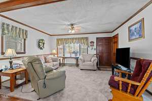 A living room with beige carpeting features two armchairs, a sofa, and a rocking chair. There's a ceiling fan, a lamp on a side table, a window with striped valances, a wreath, and a wall-mounted TV. A photo is on the opposite wall above the sofa.