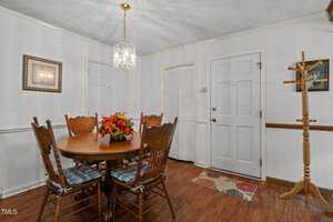 Dining room with a round wooden table and six chairs with plaid cushions. A floral centerpiece is on the table. The room features wood flooring, white walls, a chandelier, and two doors. A framed picture and a coat rack complete the space.