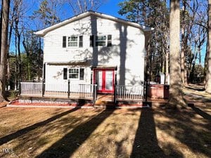A two-story white house with a red front door and multiple windows, featuring a wooden deck in front. The surrounding area has tall trees, and the ground is covered with brown grass. Shadows of the trees are cast on the house.