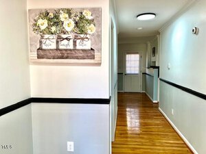 A hallway with wooden floorboards and white walls features a large floral painting on the left and a white door at the far end. The walls have a black trim, and a round ceiling light is visible.