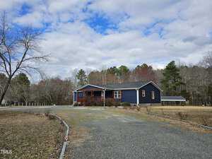 Nestled on 2126 Victory Lane, this charming single-story blue house in Creedmoor features a cozy covered porch by a gravel driveway. A lush lawn with bare trees encircles the home, while the partly cloudy sky sets a serene backdrop against a distant line of trees.